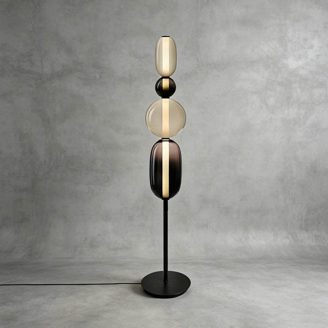 Tall Candied Model B Black glass floor lamp glowing against a textured gray studio wall.