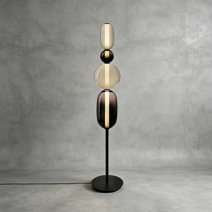 Tall Candied Model B Black glass floor lamp glowing against a textured gray studio wall.