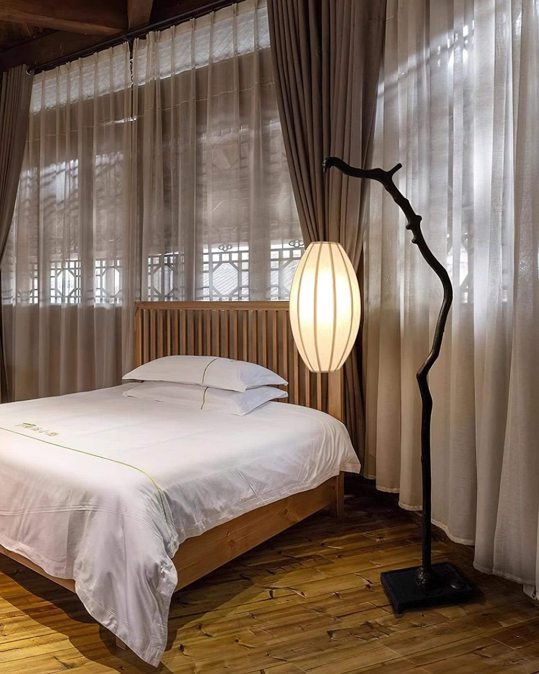 Tree floor lamp glowing beside a bed and curtains in a bedroom.