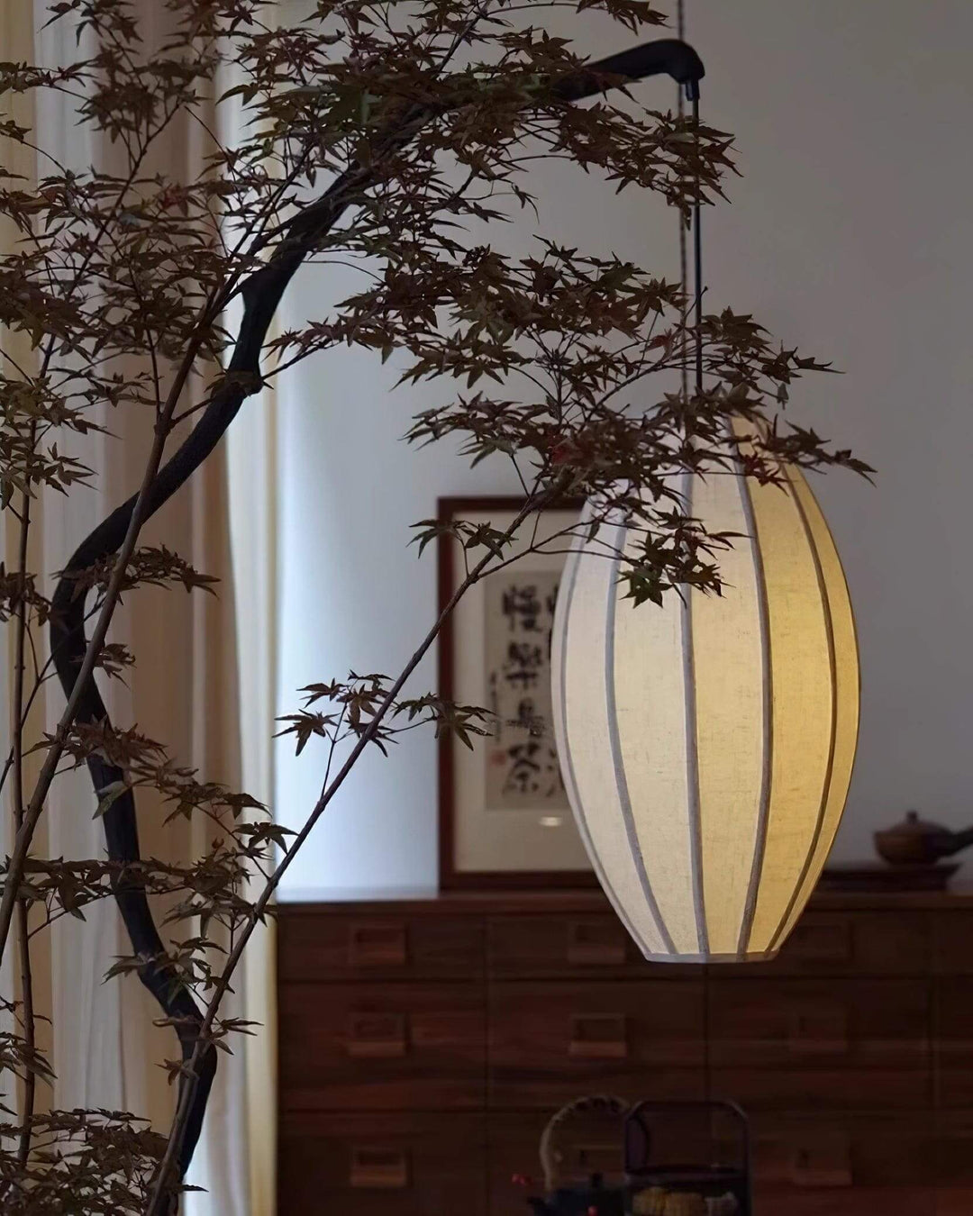 Close scene of the oval fabric shade beside a dresser and indoor tree branches.